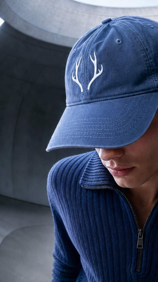 A man wearing a navy blue baseball cap with white antler embroidery and a matching navy blue ribbed quarter-zip sweater, photographed from a low angle inside a circular structure.