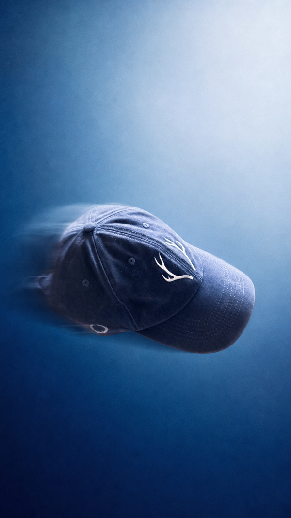 A dark navy blue baseball cap with a white embroidered deer head logo, shown floating against a blue gradient background with motion blur.